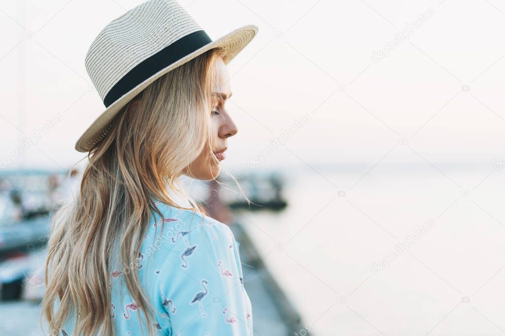 Beautiful blonde young woman in blue dress and straw hat on pier on sunset