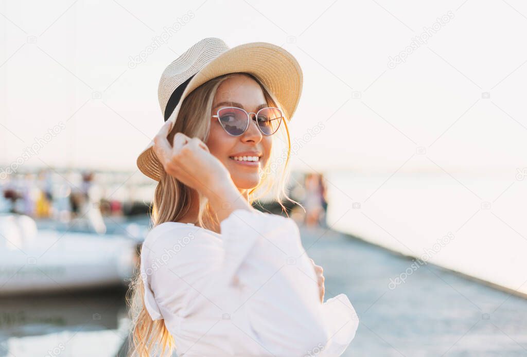 Beautiful blonde young woman in white shirt and straw hat on pier on sunset