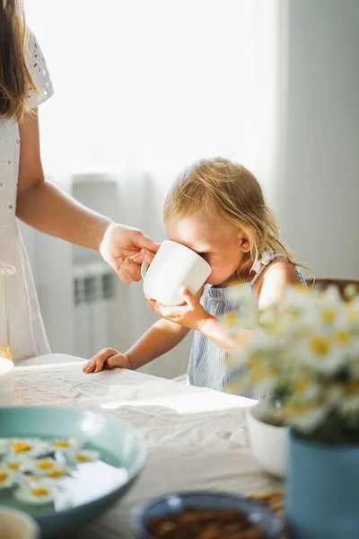 Cute blonde toddler girl drink from cup from mother\'s hand at table on sunny day. Cottagecore Aesthetics