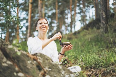 Genç, gülümseyen bir kadın ormanda Tibet müzik kasesinde oynuyor. Ses terapisi ve meditasyon