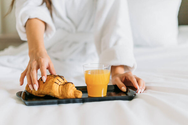 Young woman in white robe having breakfast orange juice and croissant on bed in hotel room