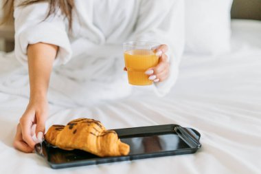 Beyaz cüppeli genç bir kadın otel odasındaki yatakta portakal suyu ve kruvasan yiyor.