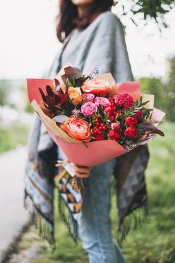 Pançolu ve kotlu genç esmer kadın elinde kırmızı tonlarda farklı çiçeklerden güzel bir buket tutuyor.