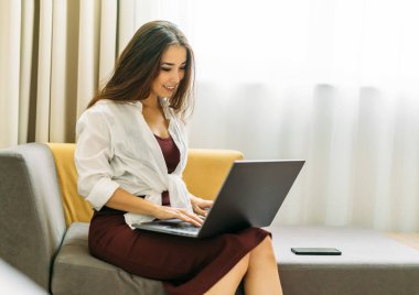 Güzel, gülümseyen Asyalı, uzun saçlı bir kadın otel odasında dizüstü bilgisayarla çalışıyor.