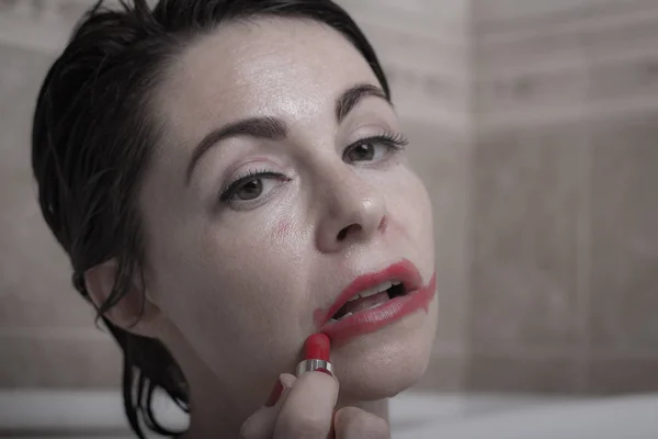 female depression. a woman in the bathroom with a petrified look slashes lipstick on her face.