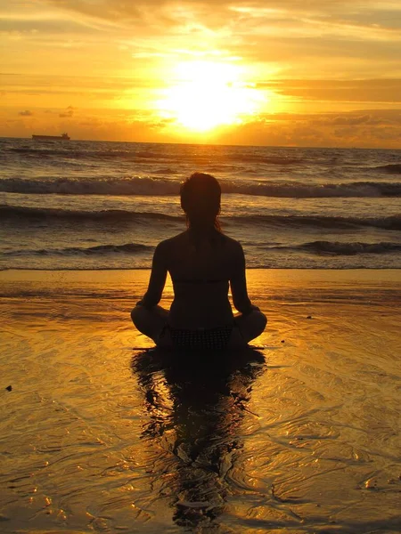若い女性は、ビーチに座って、日没のシルエット。ヨガを屋外で練習している若い女性。調和と瞑想の概念。健康的なライフスタイル蓮のヨガで瞑想し — ストック写真