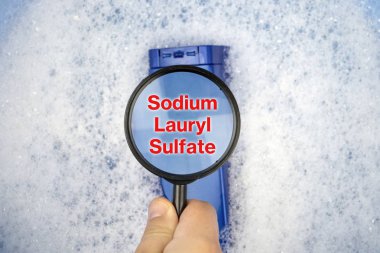 Chemical components on the shampoo label:  Sodium Lauryl Sulfate (sls, sles). A hand holds a blue jar and a magnifier, where the harmful ingredients of a detergent are written in close up. clipart