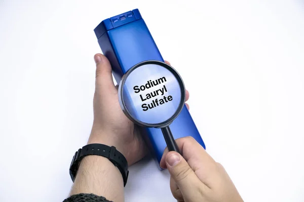 Chemical components on the shampoo label:  Sodium Lauryl Sulfate (sls, sles). A hand holds a blue jar and a magnifier, where the harmful ingredients of a detergent are written in close up.