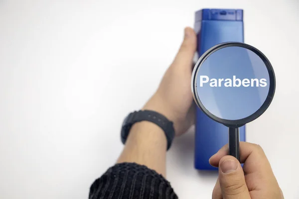 Chemical components on the shampoo label: parabens . A hand holds a blue jar and a magnifier, where the harmful ingredients of a detergent are written in close up.