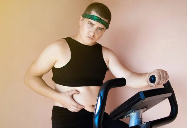 Cheerful fat man in a funny tracksuit is engaged in a stationary bike. The concept of problems with weight, losing weight, playing sports and motivation to achieve a slim body.