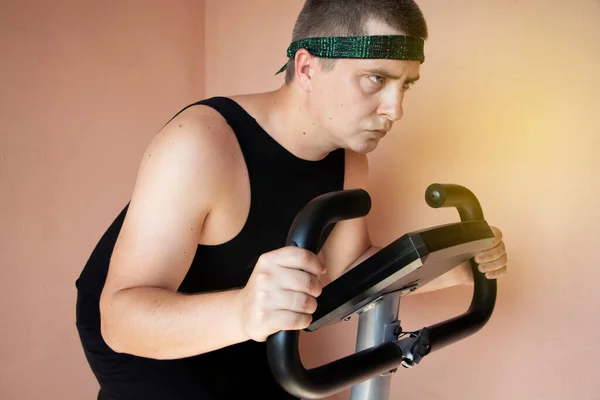 Cheerful fat man in a funny tracksuit is engaged in a stationary bike. The concept of problems with weight, losing weight, playing sports and motivation to achieve a slim body.