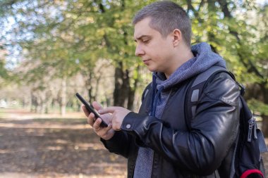 Deri ceketli bir adam ormanın ortasında telefona bakar. Sosyal medya daldırma, yönlendirme ve telefon stresi kavramı