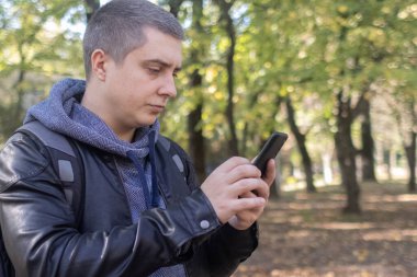 Deri ceketli bir adam ormanın ortasında telefona bakar. Sosyal medya daldırma, yönlendirme ve telefon stresi kavramı