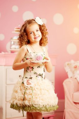 Little cute girl in a pink dress drinks tea with sweets in the children's room. Baby with curls hairstyle play in the nursery. Positive child and tea party
