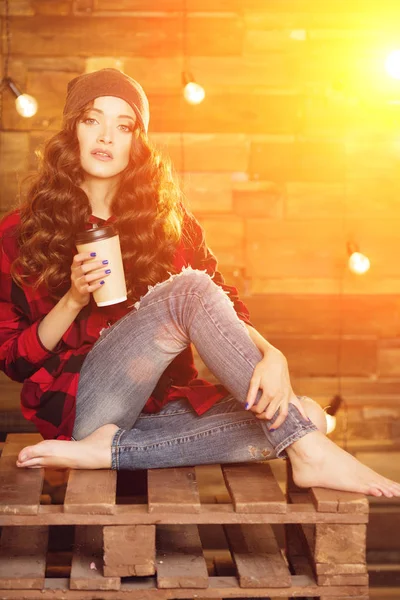 美しいモダンなファッショナブルな少女赤いドレスと破れたジーンズ ケージ内でスマート フォンと日光のライトと木製の背景にコーヒー 帽子をかぶって — ストック写真