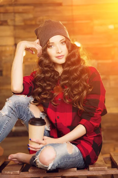 美しいモダンなファッショナブルな少女赤いドレスと破れたジーンズ ケージ内でスマート フォンと日光のライトと木製の背景にコーヒー 帽子をかぶって — ストック写真