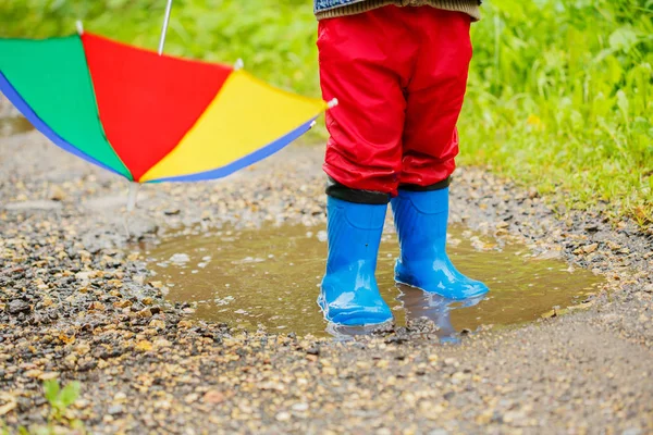 子供は ブーツに水たまりにジャンプします 雨の中での赤ちゃん 虹の傘を持つ少年は 外を歩いています — ストック写真