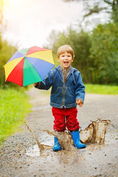 子供は ブーツに水たまりにジャンプします 雨の中での赤ちゃん 虹の傘を持つ少年は 外を歩いています — ストック写真