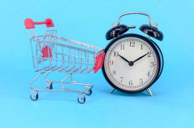 Alışveriş arabası ve mavi arka planda klasik çalar saat. Satış zamanı market tüketicisi konsepti. Seçici odak.