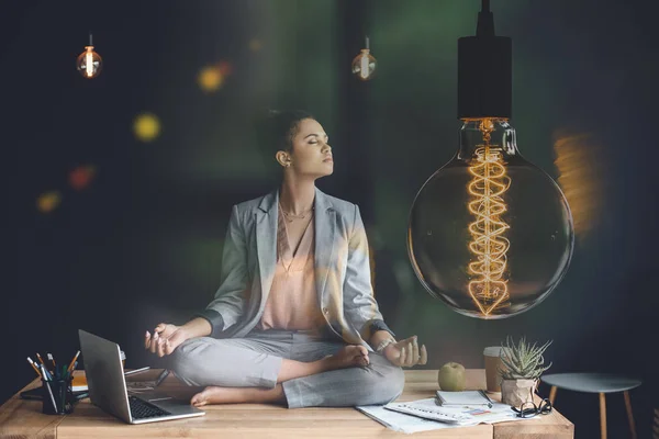 Light bulb representing creative thinking and young businesswoman meditating in lotus position on work table — Stock Photo