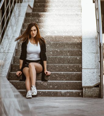 Kızıl saçlı güzel bir kız bir siyah hırka ve kısa siyah şort giyiyor. Gri kentsel basamaklarında oturan kamera poz.