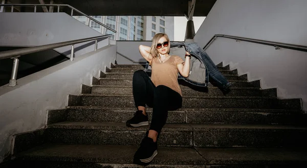 Блондинка в солнечных очках одета в джинсовую куртку, сидящую на ступеньках. Современная девушка — стоковое фото