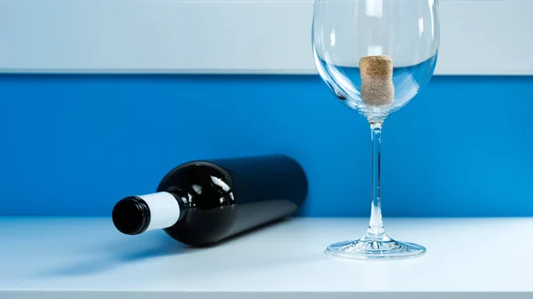 Vacíe la copa de vino en una mesa blanca junto a una botella de vino vacía de vidrio oscuro. Corcho dentro — Foto de Stock