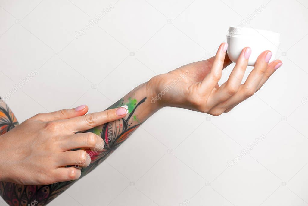 Female tattooed hands hold a jar of cream. Fingers apply cream to skin