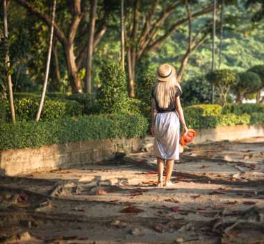Bir hasır şapka ve uzun bir etek Sarışın tropikal bir parkta yürür. Geri görünüm. Tayland seyahat