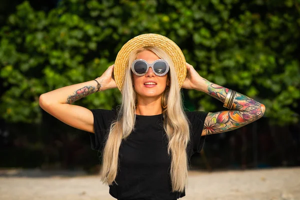 Vacker blondin ler och tittar på morgon havet. Håller en hatt med händerna med tatueringar. Resa till Thailand. — Stockfoto