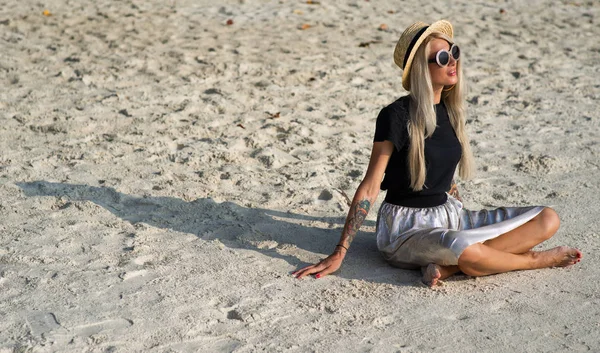 En flicka i en stråhatt sitter på morgon stranden. Modern flicka på semester — Stockfoto