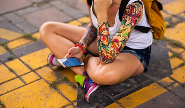A modern lány egy útjelzésen ül. Hordoz egy sárga hátizsák. Tetovált kezek — Stock Fotó