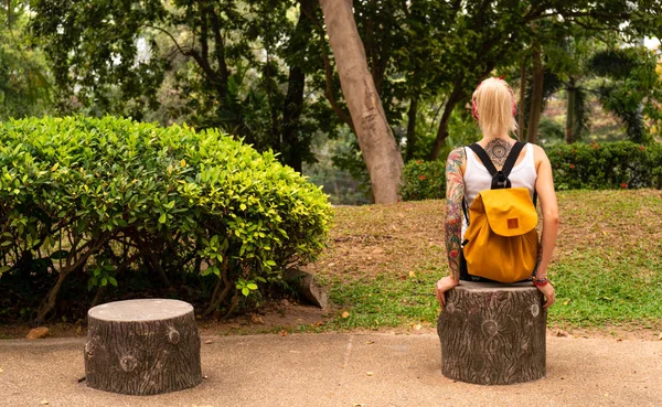 O blondă cu un rucsac galben stă într-un parc verde. Poartă un bandaj roşu, braţe cu tatuaje. Asia călătorie — Fotografie, imagine de stoc