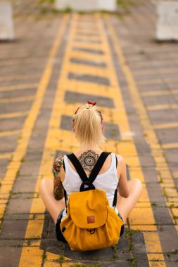 Modern kız bir yol işaretleme üzerinde kameraya sırtını oturur. Sarı bir sırt çantası, silah dövmesi taşır