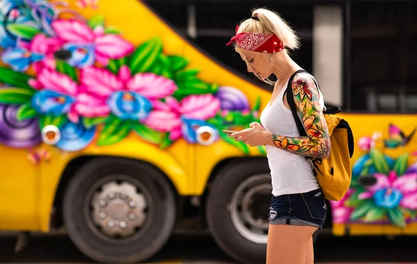 Een blonde vrouw met een telefoon in de hand staat tegen de achtergrond van een gele bus. Modern meisje draagt een rode bandage, armen getatoeëerd. Stockfoto