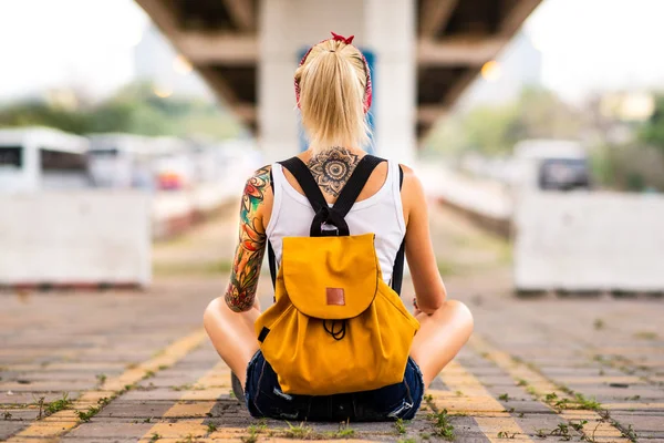 Современная девушка сидит спиной к камере на дорожной разметке. С желтым рюкзаком, татуировкой на руках. — стоковое фото