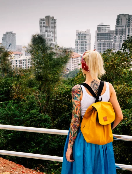 De blonde staat met zijn rug naar de camera, kijkend naar de stad vanaf een hoogtepunt. Het dragen van witte trui, blauwe rok, oranje rugzak. Handen met tatoeages. Asia Travel Stockafbeelding