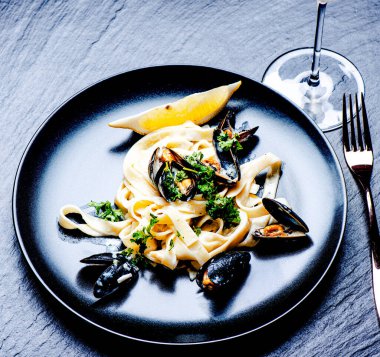 Taş yüzeyinde siyah tabakta mavi midye ve makarna.