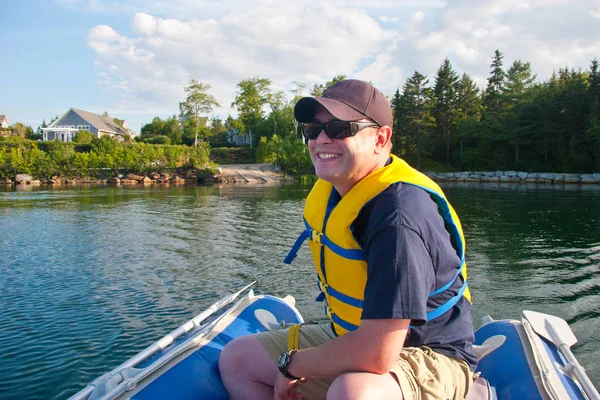 A man wearing a hat and a lifejacket grins with glee as he drives a small blue dinghy around a lake or sea.