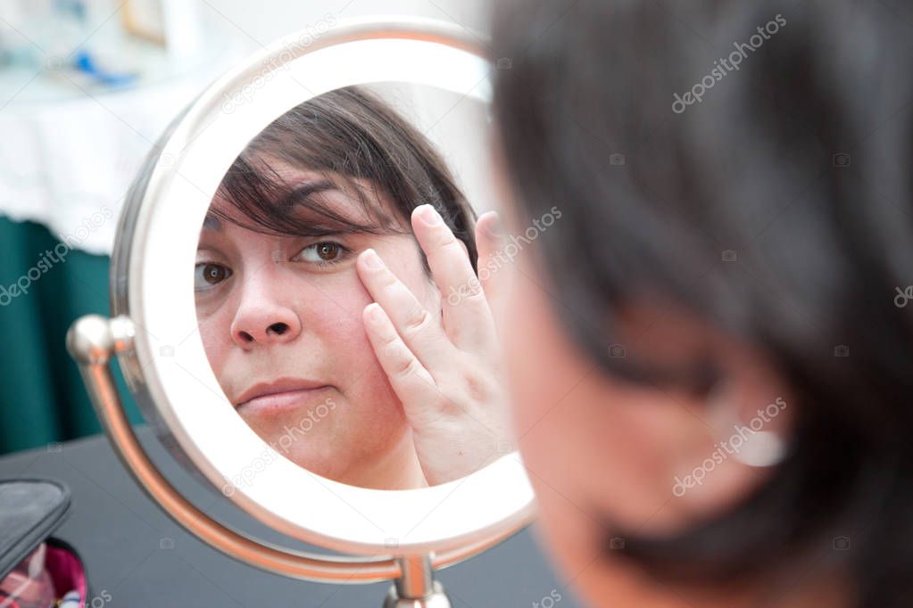 A woman looks at herself in a mirror and looks at her pores and skin, touching her eye 