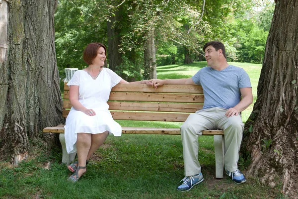 A happily married man and woman sit together on a park bench and look affectionately at one another