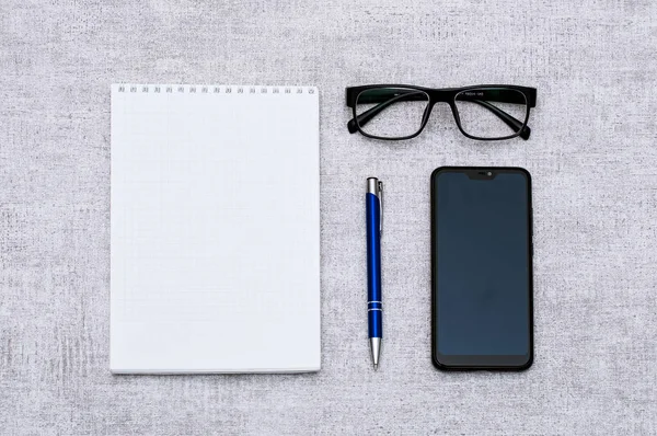 Blank notepad with blue pen andsmartphone and glasses flat lay. Top view on  stationery with mobile phone and spectacles. Art, business, creation, imagination, office supplies and business concept.