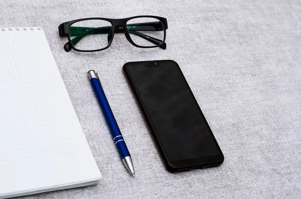 Blank notepad with blue pen and smartphone and glasses flat lay. View on stationery with mobile phone and spectacles. Art, business, creation, imagination, office supplies and business concept. Contain empty space for your design or montage.