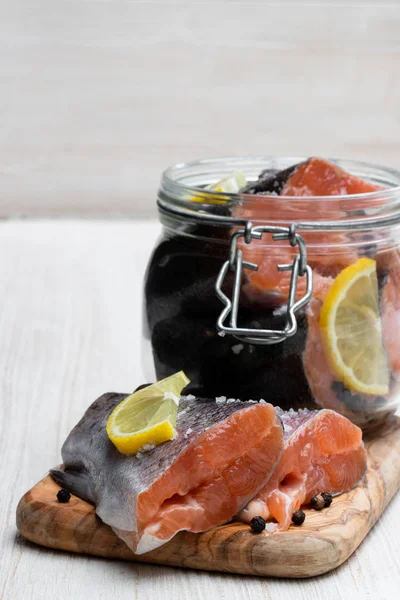 Salted rainbow trout slices with lemon in glass jar on white wooden table