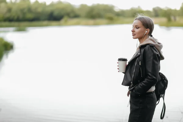 Una hermosa mujer en una chaqueta de cuero camina por el parque con auriculares y bebe café junto al lago . —  Fotos de Stock