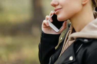 Deri ceketli güzel bir kadın parkta yürüyor ve telefonda konuşuyor. Yakın plan planı.