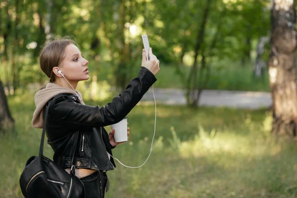 Una hermosa mujer en una chaqueta de cuero y auriculares camina por el parque, escucha música y se toma una selfie . — Foto de Stock