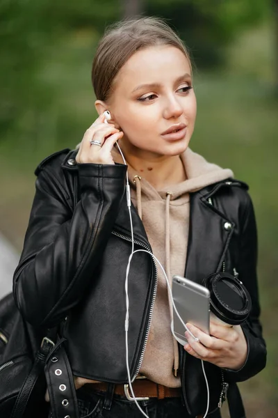 Una hermosa mujer en una chaqueta de cuero camina por el parque, inserta auriculares en sus oídos para escuchar música — Foto de Stock