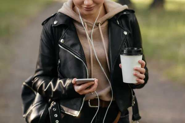 En kvinna med hörlurar i öronen håller telefonen i handen och tittar in i den. Går i parken. — Stockfoto