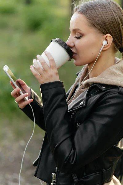 Una hermosa mujer con auriculares camina por el parque, habla en una videollamada desde un teléfono y bebe café . — Foto de Stock